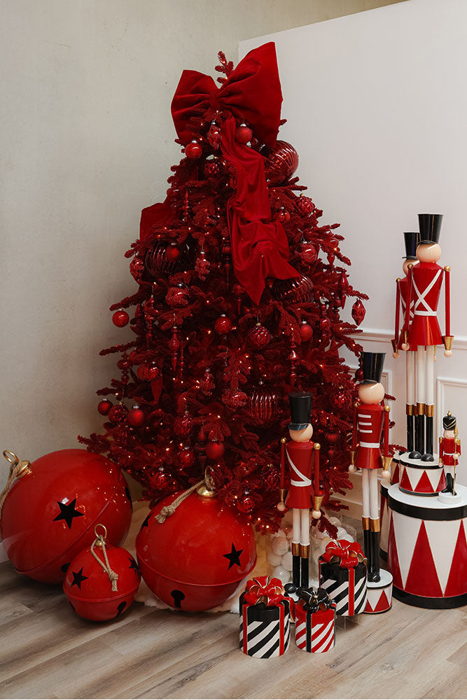 Pre Lit Ruby Red Christmas tree with red ornaments and large bow, surrounded by festive decorations on a wooden floor.