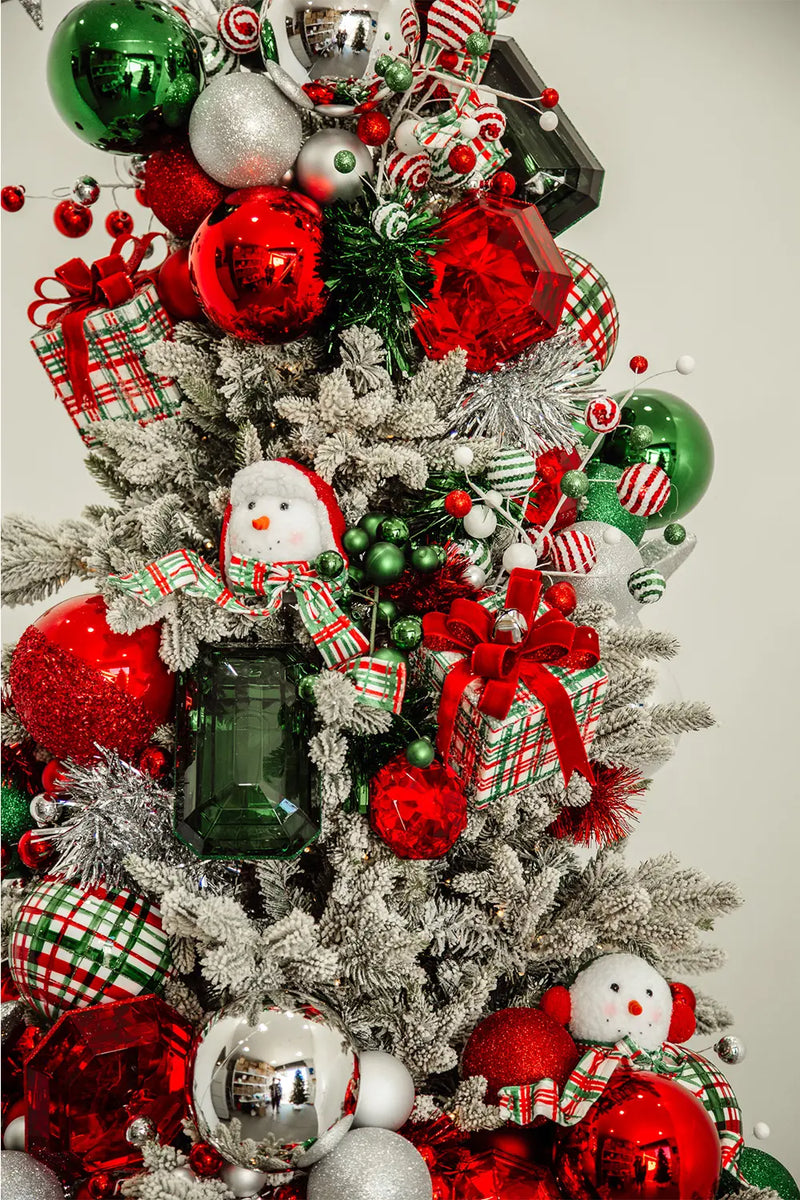 A close-up of a Christmas tree, displaying a dense arrangement of ornaments in colors predominantly red, green, and silver. Notable decorations include a snowman with a scarf, glossy red and green balls, and candy cane inspired stripped ornaments..
