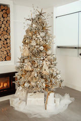 An elegant Christmas tree adorned with gold and silver ornaments, standing next to a fireplace with neatly stacked logs. The tree is decorated with starbursts, angels, and various sparkling ornaments. Below the tree are white gift boxes wrapped with satin bows. The background features a white room with a glass door.