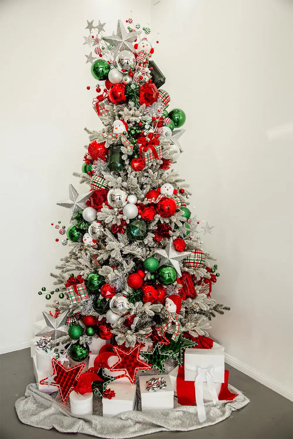 A Christmas tree, richly decorated with green, silver, and red ornaments. It's adorned with a variety of items including snowmen, stars, and wrapped presents placed beneath it. A silver blanket lies underneath.