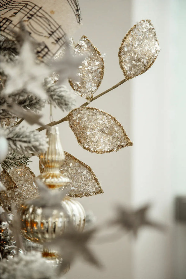 Close-up of shimmering leaf-like decorations on the Christmas tree. These glittering ornaments have a crystal-like texture, edged in gold, reflecting the ambient light. In the background, blurred silvery ornaments and pine branches can be seen.