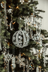 Close-up view of a Christmas tree showcasing intricately designed sparkling ornaments, including a captivating silver cage-like bauble with embedded jewels, alongside hanging crystal droplets, and other shimmering decorations, bathed in the warm glow of tree lights.