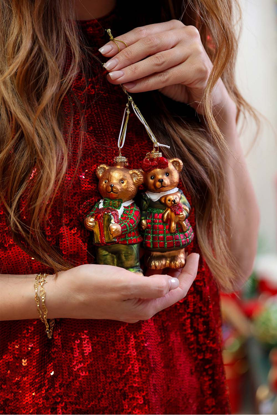 Hands holding a pair of teddy bear Christmas ornaments against a red sequin dress